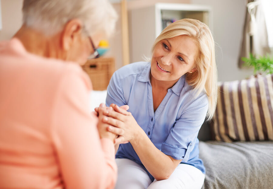 Smiling caregiver holding hands with a senior woman in a cozy living space.