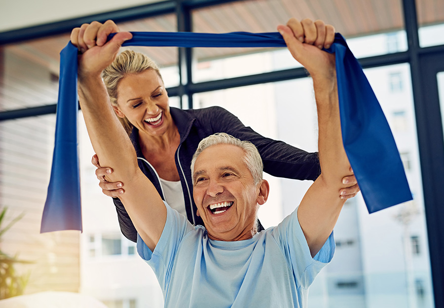 Instructor assisting elderly man with exercise band indoors, both smiling.