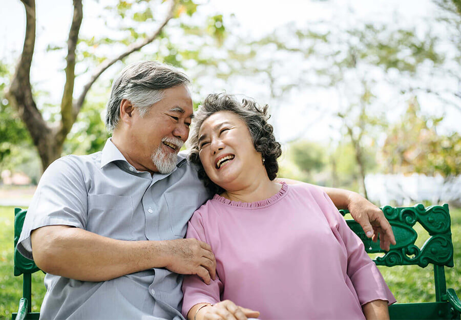 Elderly couple sitting on a bench, laughing and enjoying time in a green garden.