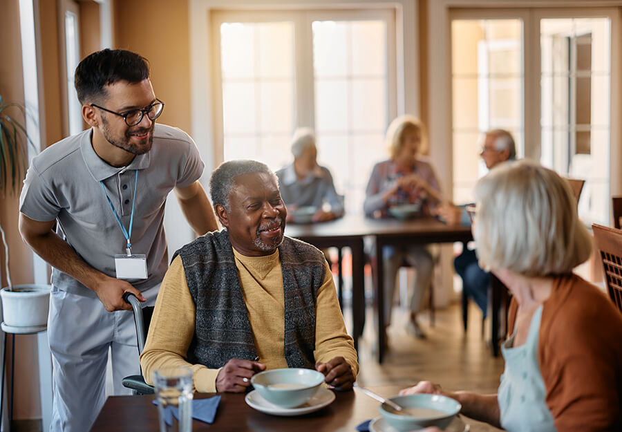 Smiling senior living caregiver assists senior man in wheelchair going to dinner.