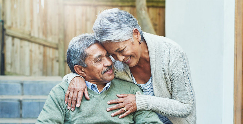 Senior couple embracing outdoors, man in wheelchair and woman leaning in affectionately.