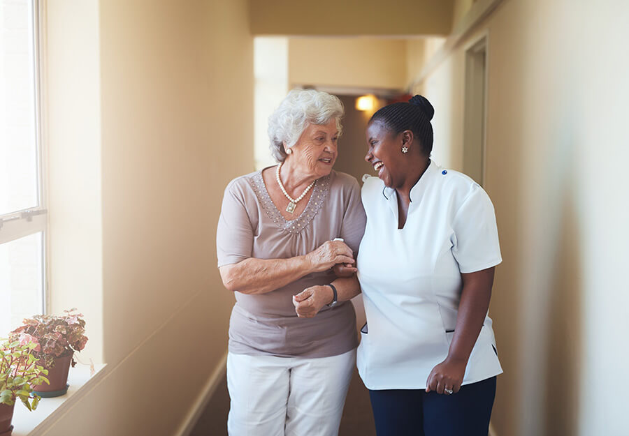 Senior woman and nurse smiling while walking in the hallway of senior living community.