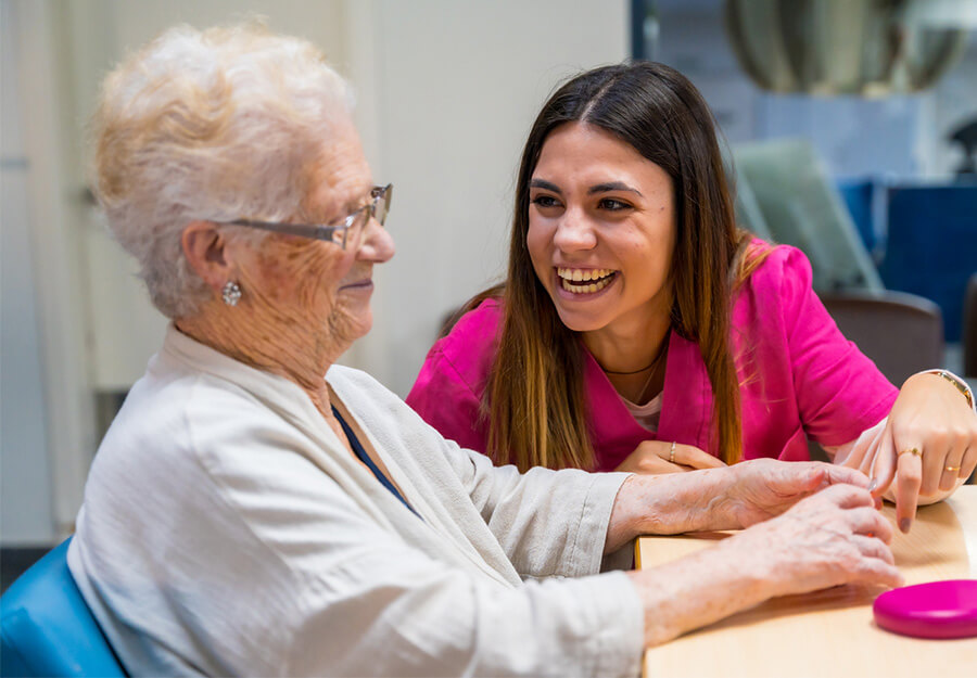 Senior living caregiver laughs with elderly woman at a table.