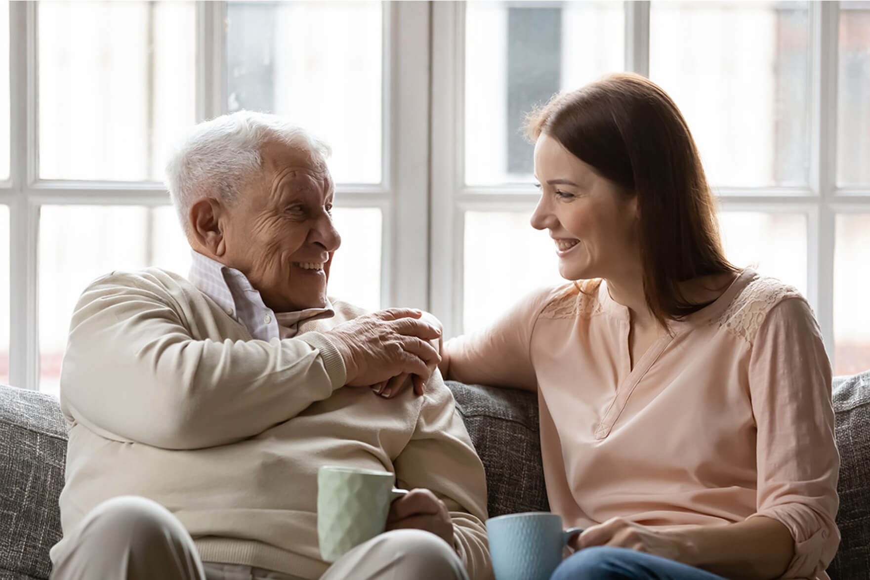 Elderly man and young woman smiling while sitting on a sofa, holding mugs.