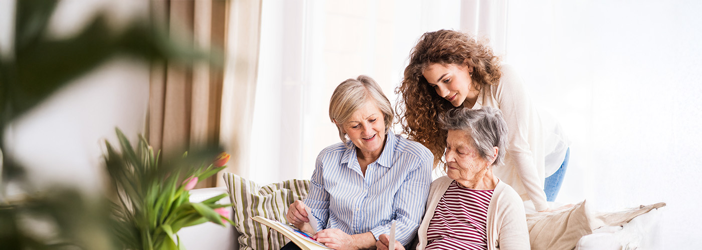 Two senior women sit on couch and review a photo album while younger woman looks on